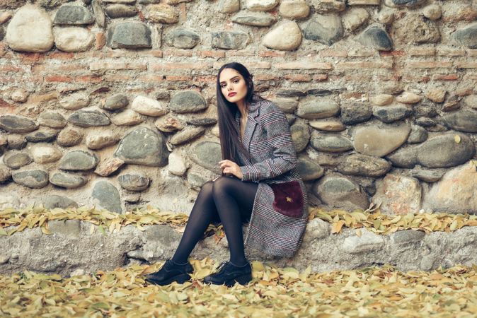 Female looking at camera in warm winter clothes sitting in front of rock wall in autumn time