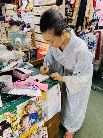 Japanese older woman in kimono writing on a small notebook in a store shop