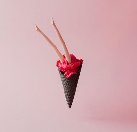 Doll legs emerging from strawberry ice cream in dark waffle cone on pink pastel background