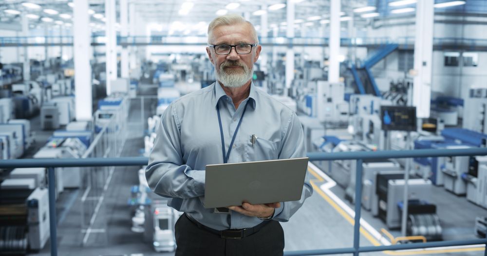Portrait of Engineer Wearing Glasses, Using Laptop Computer and Looking at Camera. Technician in High-VIsibility Vest Working in a Modern Technology Factory
