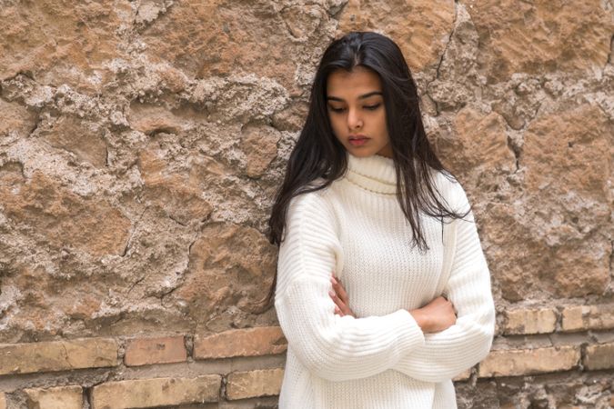 Sad young woman in light turtleneck sweater standing beside brown rock