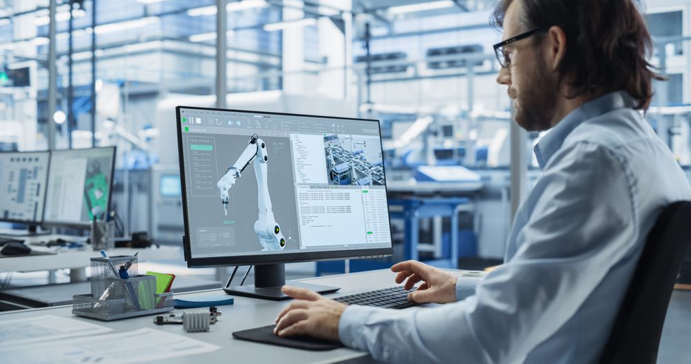 System Administrator Working on a Desktop Computer at a Modern Manufacturing Plant. Software Developer Programming the Work of the Robotic Arms, Monitoring Production on the Assembly Conveyor