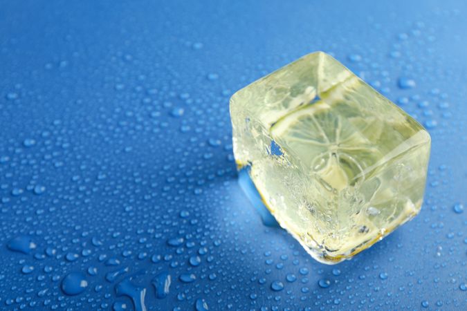 Top view of large rectangular ice cube with lemon slice on blue table, copy space