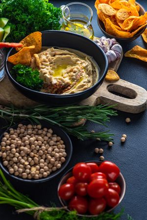 Creamy hummus dip in bowl on board served with olive oil, veggies, chick peas and cherry tomatoes