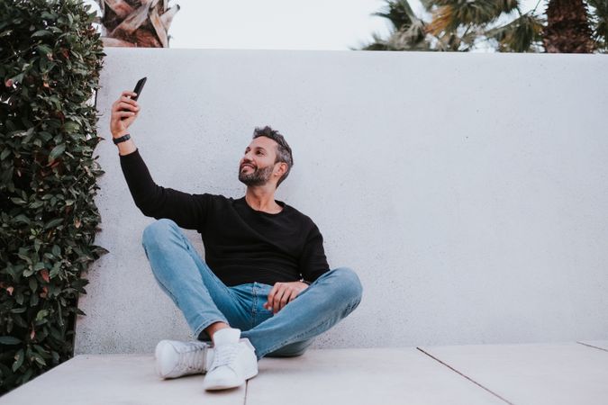 Adult stylish smiling man taking selfie on smartphone near grey wall