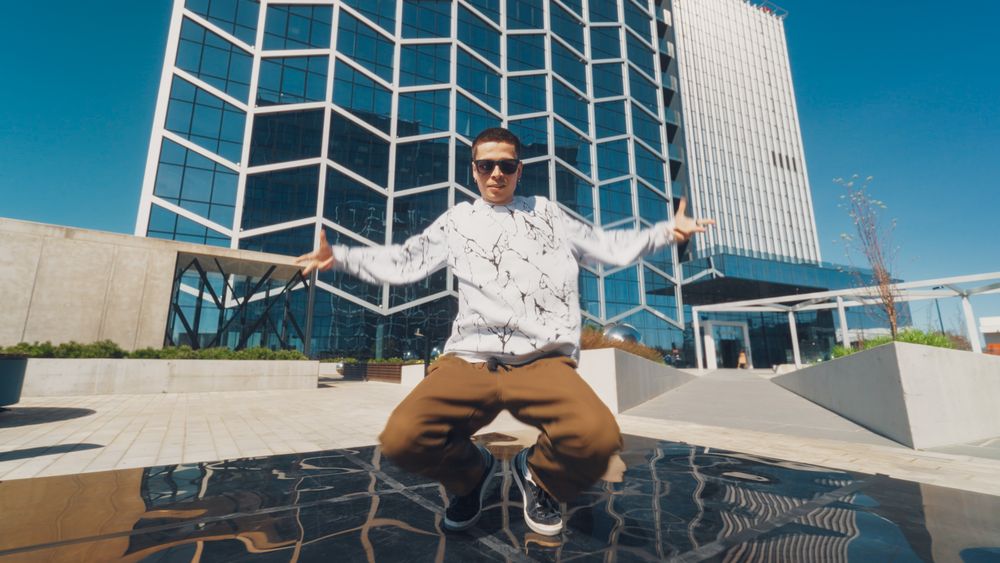 A Young Man Showcases His Breakdancing Skills In Front Of A Modern Glass Building, Under A Clear Blue Sky.