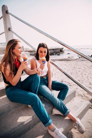 Two young women enjoying ice cream cones while sitting on the steps of a beach entrance