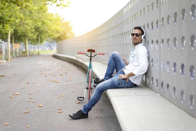 Male in sunglasses sitting outside of park listening to music with bicycle