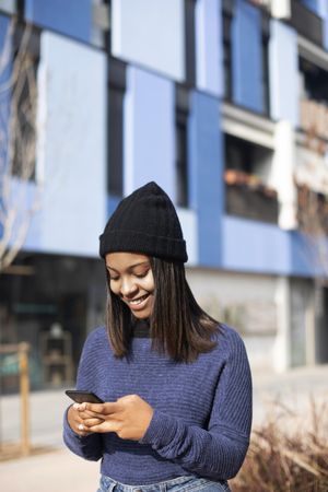 Female in hat and sweater checking cell phone outside blue building, vertical
