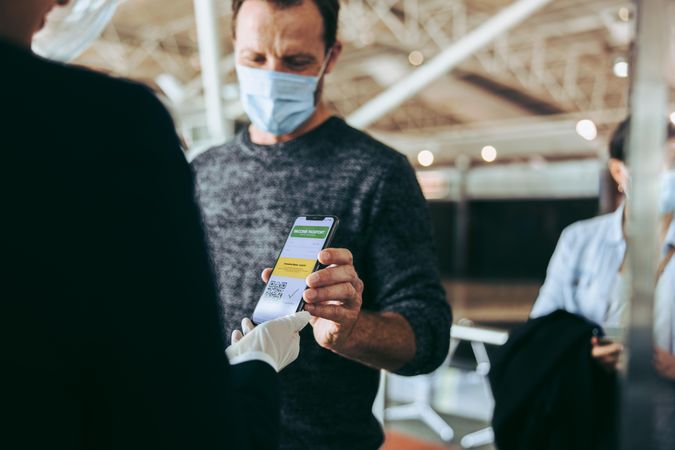 Man in face mask showing his covid-19 vaccine passport on cell phone to airport staff