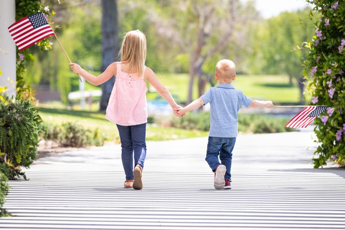 Young Sister and Brother Holding Hands and Waving American Flags At The Park