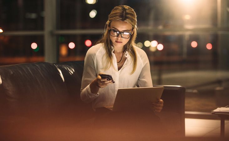 Young businesswoman texting on mobile phone while working on laptop late in office