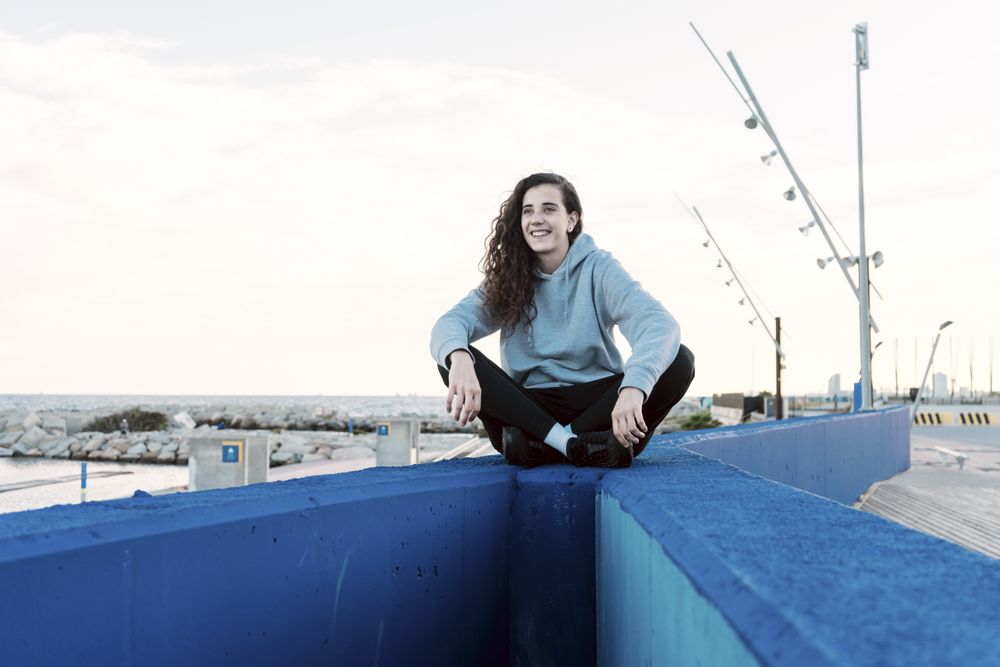 Young teenage girl sitting on a stone wall with her legs crossed while smiling at the coast
