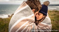 Young couple embracing under blanket in a chilly day with sea and dark cloudy sky in the background 4joYxb