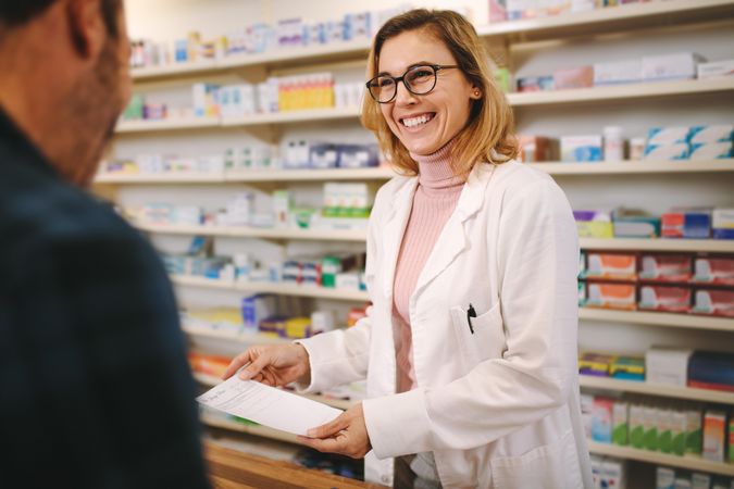Smiling female pharmacist with prescription assisting a customer standing at the counter