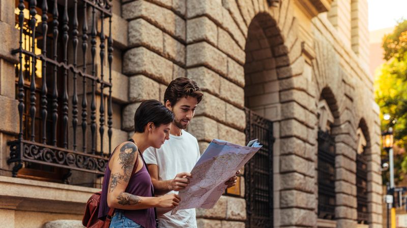 Man and woman travelers standing near an old stone building with a map
