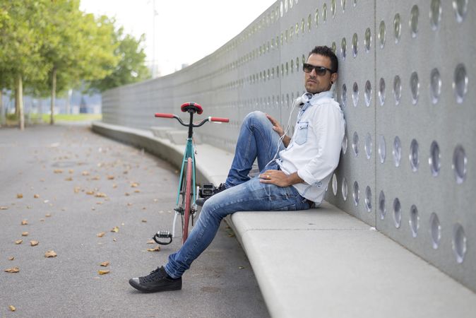 Male in sunglasses relaxing outside of park with headphones and bicycle