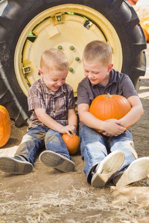 Two Boys Holding Pumpkins Talking and Sitting Against Tractor Tire