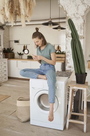 Relaxed women sitting on washing machine and browsing smartphone