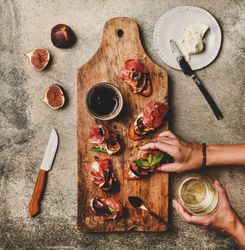 Hands with canapé of prosciutto, goat cheese and grilled figs, white wine, square crop