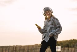 Happy young woman using smartphone and listening to music in headphones in countryside