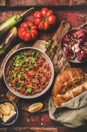 Eggplant dip with pomegranate and parsley garnish on wooden surface with vegetables and bread