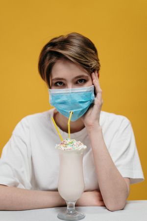 Blonde person with facemask sitting at a table with smoothies against yellow background