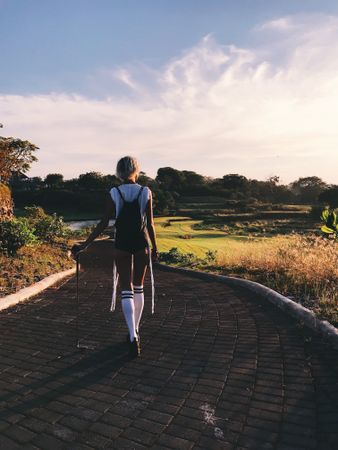 Back view of young woman walking on asphalt between grass fields at sunset