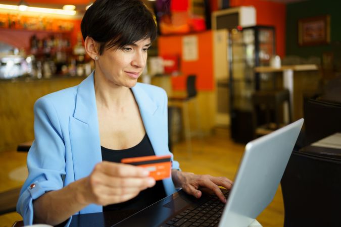 Female in trendy blue jacket sitting in cafe shopping on laptop with credit card