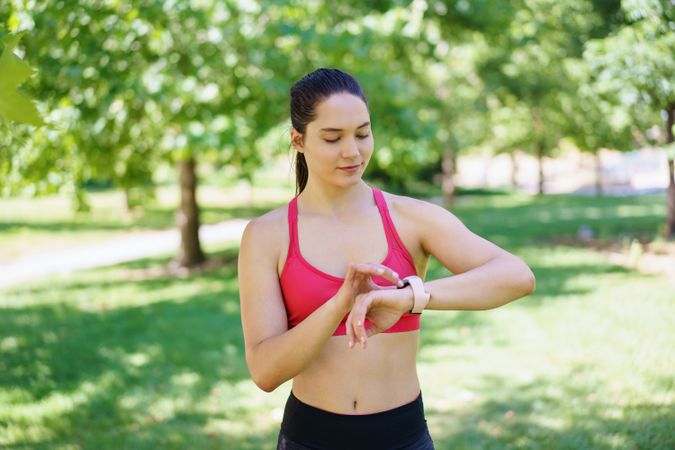 A young woman in a pink sports bra checks her fitness tracker in a park, focused on her workout routine