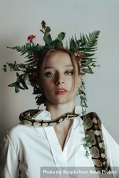 Young woman in light top with floral headdress and live snake on her shoulder against light background 0LWoDb