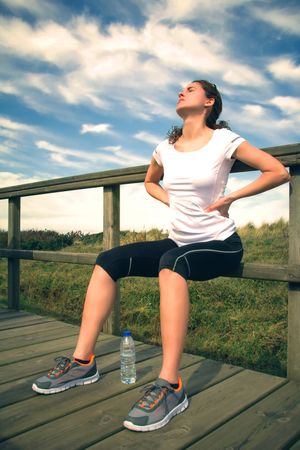 Woman in athletic gear holding hurt back while seated on wooden path