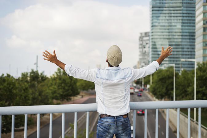 Male in denim standing with open arms on bridge over city traffic