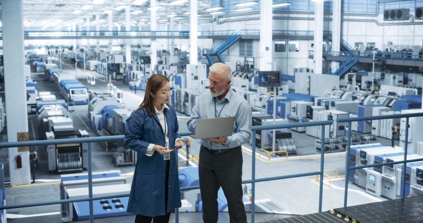 Two Multiethnic Engineers Standing on a Platform, Using Laptop Computer at an Electronics Factory. Machines are Undergoing Maintenance, Specialists Monitoring the Progress Through Online Software