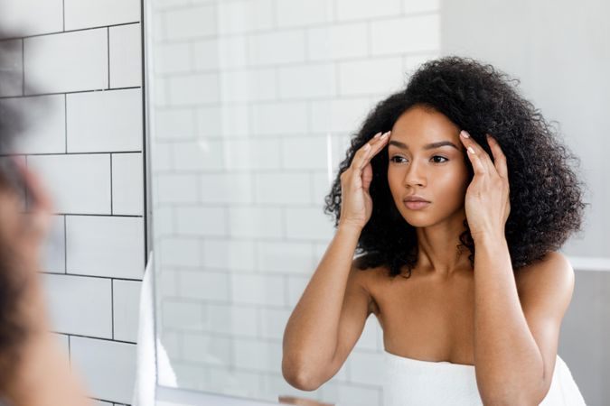 Black woman with curly hair touching her temples in the mirror