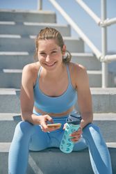 Happy sportswoman with smartphone and bottle resting on stairs o