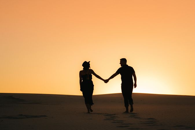 Silhouette of man and woman holding hands standing on sand during sunset