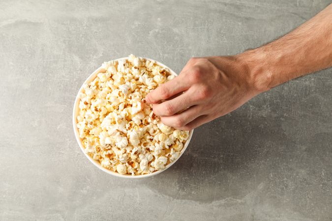 Male hand and bucket with popcorn on grey background, top view