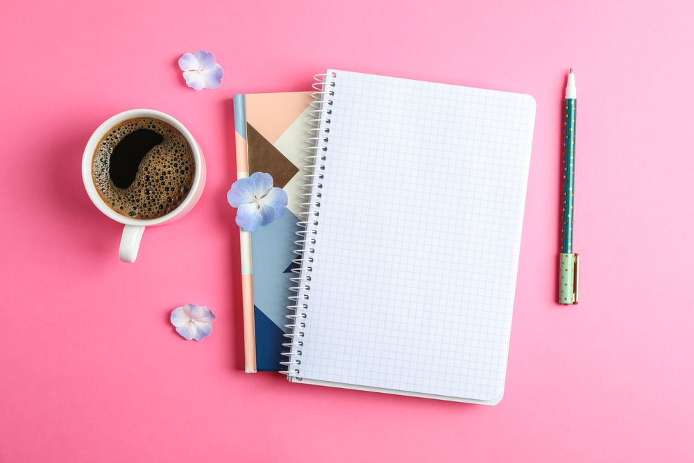 Cup of coffee with petals of flower, copybooks and pen on color background, top view