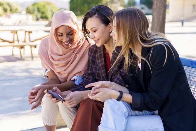 Smiling women sitting on outdoor park bench talking while watching smartphone