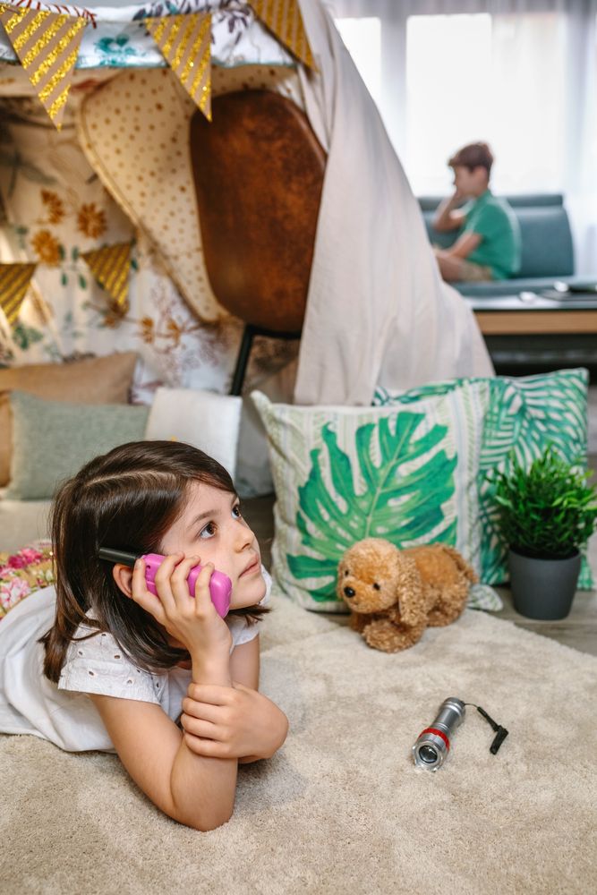 Pensive little girl holding walkie-talkie lying over rug in tent while talk with boy sitting on sofa