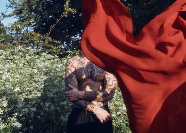 Tattooed male standing in garden with red sheet hiding his face