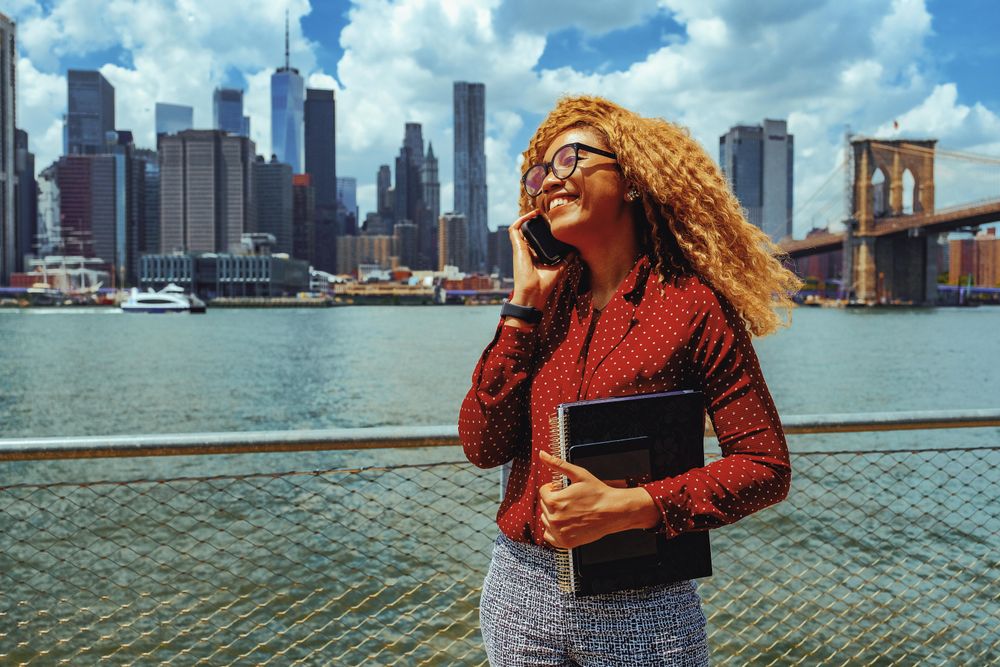 Smiling professional Black female speaking on phone while holding her notebooks by Hudson River