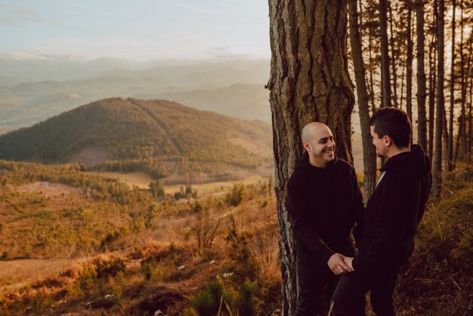 Smiling men hugging near tree in forest on hill