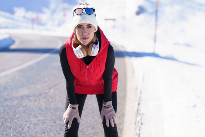 Determined fit woman in warm clothes and headphones leaning on knees while looking at camera wintry road