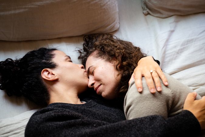 Two women lying down embracing on bed, one kissing the other’s forehead