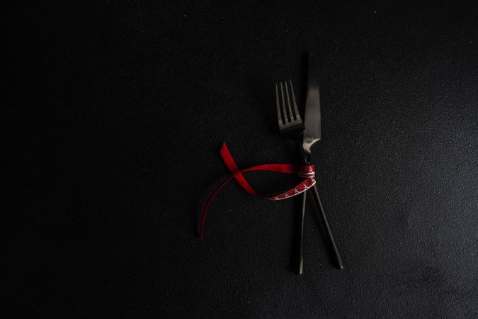 Dark knife and fork on dark background tied with bright red ribbon