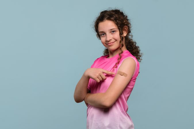 Smiling girl with bandage on arm pointing at it.