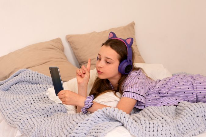 Girl using smartphone in bed with headphones, focused expression.