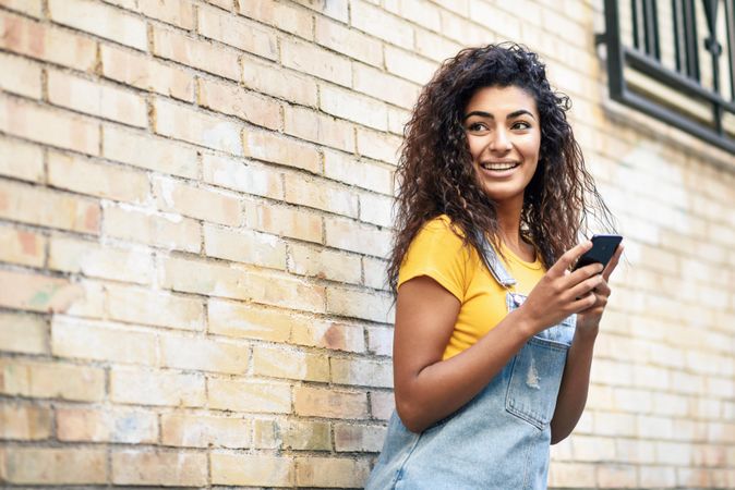 Arab woman looking around while texting with her smart phone outside in front of brick wall